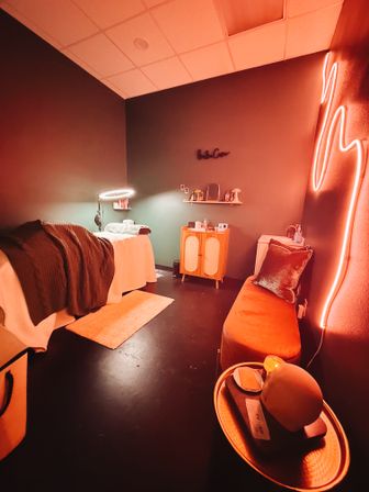 Cozy spa treatment room with massage table and blanket, illuminated ring light, neon pink wall accent, velvet bench and mid-century cabinet bathed in warm orange glow