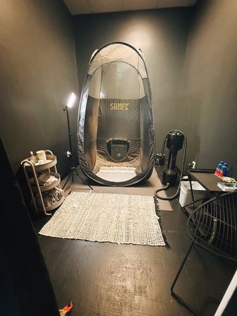 Private indoor tanning room with a pop-up spray-tan tent, tripod lamp, extraction fan/spray machine, towel cart, woven rug and a side chair — compact salon setup.