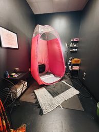 Pink pop-up spray-tan tent in a compact dark-walled salon room with rugs, supply shelves and a ring light — cozy tanning studio setup