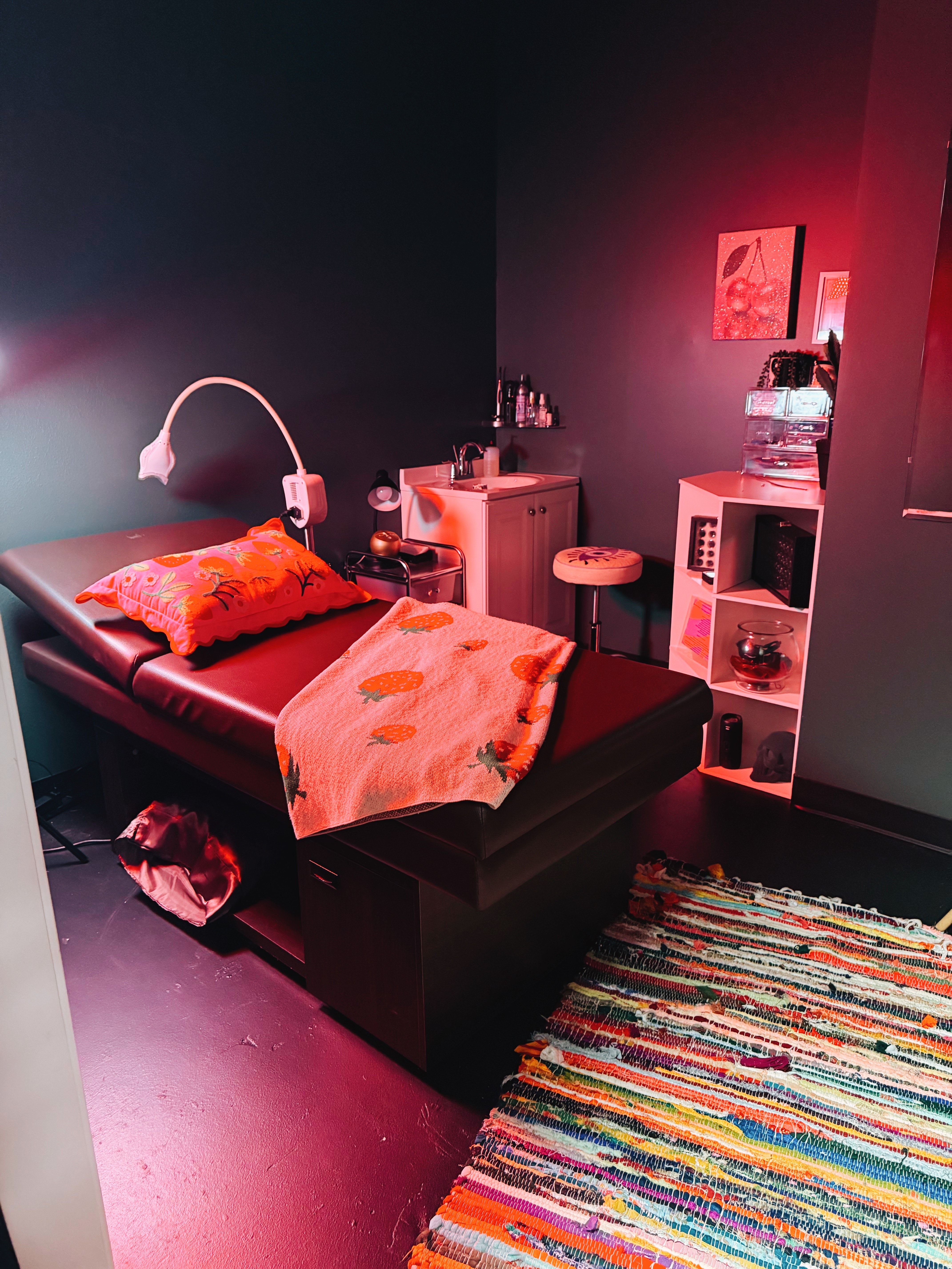 Cozy spa treatment room with dark walls, a padded massage table topped with a floral pillow and orange blanket, pink ambient lighting, small sink and storage shelving, and a colorful woven rug.