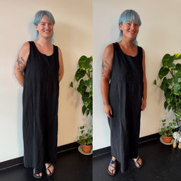 Smiling person with blue hair and tattoos wearing a black sleeveless maxi dress and sandals, standing indoors against a pale wall next to potted houseplants.
