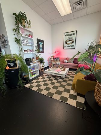 Cozy plant-filled waiting lounge with a pink sofa, mustard armchair, checkerboard rug, white coffee table and shelving, colorful decor and a 'No Pressure No Diamonds' wall poster.