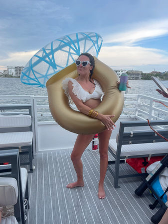 Person in a white ruffled bikini and heart-shaped sunglasses posing with a gold inflatable cocktail float and blue umbrella on a boat deck, holding a colorful tumbler with coastal condos and calm bay waters in the background.