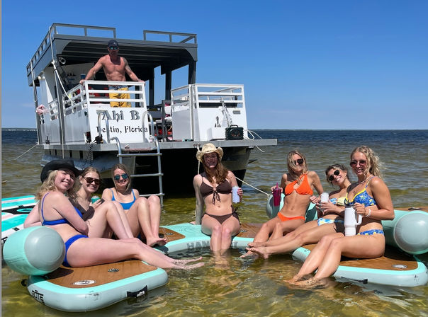 Group of friends in colorful swimsuits lounging on linked paddleboards in shallow coastal water beside a docked party pontoon boat, holding drinks and enjoying a sunny blue-sky day.