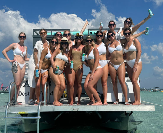 Group of women in white swimsuits posing on a pontoon boat over clear turquoise water and a sunny blue sky, holding drinks for a lively boat-party vibe.