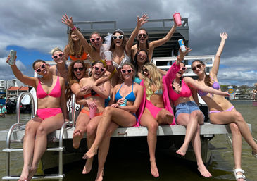 Cheerful group of women in colorful bikinis and heart-shaped sunglasses holding drinks and celebrating a bachelorette party on a pontoon boat by the shoreline under a cloudy sky