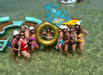 Group of women in colorful swimsuits and bucket hats posing waist-deep in crystal-clear shallow water around a giant inflatable diamond-ring float, holding drinks beside floating platforms — sunny beach party vibe.