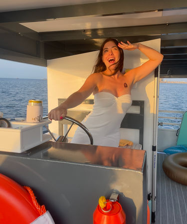 Smiling woman in a white sundress playfully steering a boat at sunset with ocean view, life ring and inflatable tube on the deck