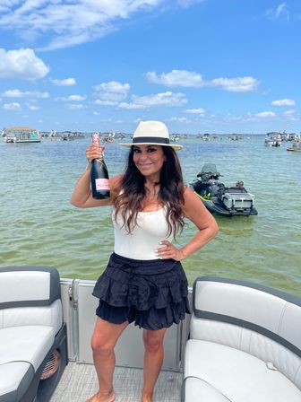 Smiling woman on a pontoon boat holding a bottle of rosé champagne, wearing a straw hat, white tank top and black ruffled skirt, with crowded boats and jet skis in shallow coastal waters under a bright blue sky with puffy clouds.