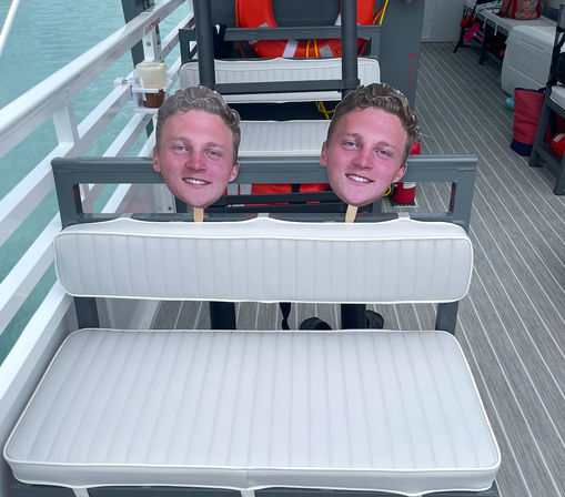 Two oversized cardboard face cutouts on wooden sticks tucked into a white padded bench aboard a pontoon boat deck, striped flooring, an orange life ring, and turquoise water visible in the background.
