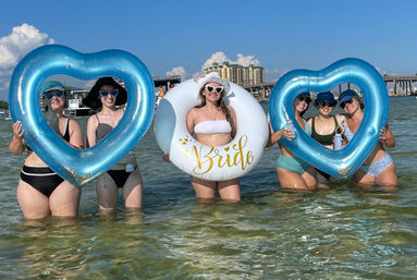 Sunny beach bachelorette party in shallow clear water: bride in a white hat inside a white float ring labeled "Bride" surrounded by bridesmaids holding blue heart-shaped inflatables, with beachfront condos and a bridge in the background.