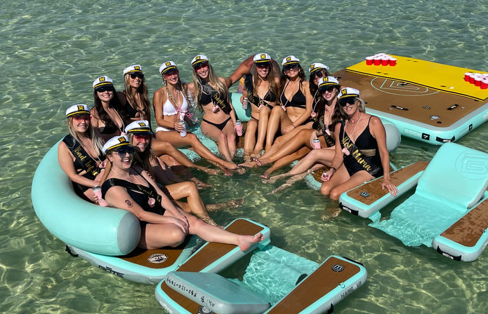 Bachelorette party on interconnected aqua inflatables in clear shallow turquoise water — group of women in captain hats and sashes, swimsuits, drinks, and a floating beer-pong board