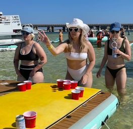 Summer beach party: women in swimsuits playing floating beer‑pong with red solo cups on an inflatable table in shallow clear water near a seaside bridge