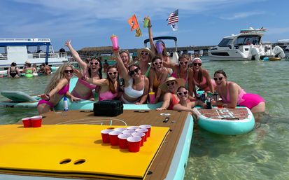 Group of women in bright swimsuits and heart-shaped sunglasses partying on paddleboards and a floating dock in shallow clear water, holding drinks around a red-cup beer-pong setup with boats and a pier in the background.