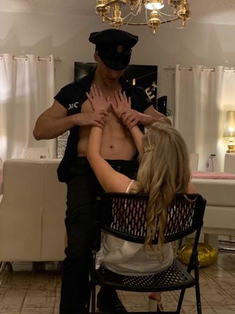 Home bachelorette party in a living room: bride-to-be with BRIDE hair clip seated while a shirtless man in a police costume performs a playful lap dance.