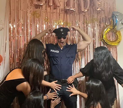 Playful bachelorette party photo: man in police costume and hat posing in front of rose-gold fringe backdrop with 'BRIDE' balloons while several women reach toward him.