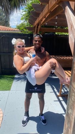 Smiling man in a baseball cap carries a woman in a white bikini and sunglasses on a sunny backyard patio under a wooden pergola, both laughing while she holds a canned drink.