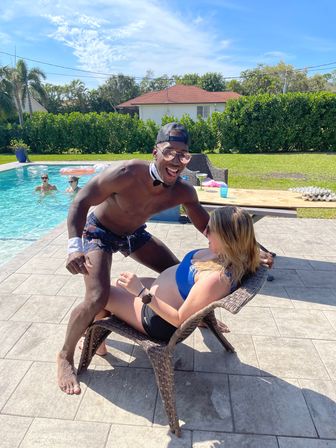 Sunny backyard pool party with a smiling man in sunglasses, a backward cap and novelty bow tie leaning over a laughing woman reclining in a wicker chair in a blue bikini top; friends float in the pool and palm trees and hedges frame the suburban yard.