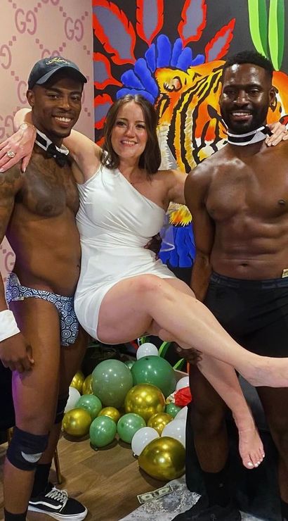 Smiling woman in a white one-shoulder dress being lifted by two shirtless male dancers wearing bow ties and cuffs, colorful tiger mural on the wall and green, gold and white party balloons on the floor.