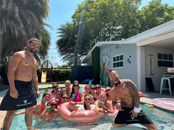 Sunny backyard pool party with a group of women on pink inflatables and two shirtless hosts by palm trees and a flamingo mural