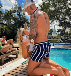 Tattooed man in striped swim trunks kneeling poolside as a woman in a white bikini holds a plastic cup, friends lounging on deck chairs under palms at a sunny backyard pool party.