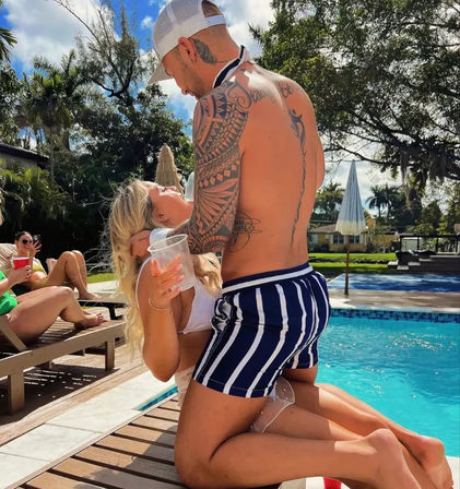 Tattooed man in striped swim trunks kneeling poolside as a woman in a white bikini holds a plastic cup, friends lounging on deck chairs under palms at a sunny backyard pool party.
