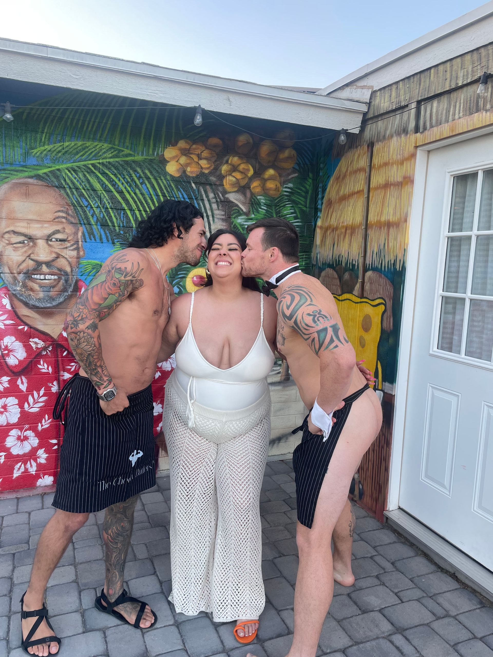 Smiling woman in a white swimsuit and crochet pants gets playful cheek kisses from two shirtless, tattooed men wearing striped aprons on a sunny paved patio in front of a colorful tropical tiki and palm mural.