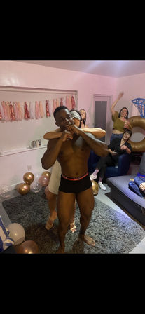 Shirtless man in black briefs playfully hugged from behind by a blindfolded woman at a lively indoor house party in a decorated living room with pink tassel garland, gold balloons, and friends cheering.