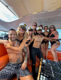 Happy group on a yacht party — friends in captain hats and black swimwear gathered around a DJ controller with ocean and blue-sky backdrop