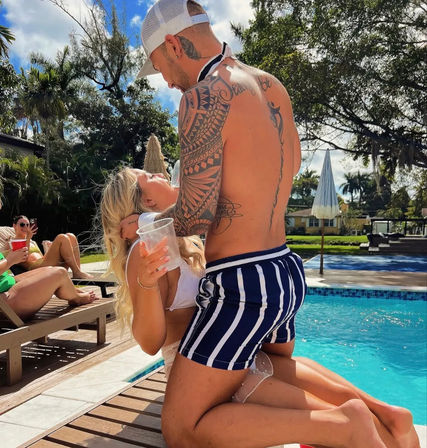 Sunny backyard pool scene: tattooed man in striped swim trunks kneels on the deck leaning toward a woman in a white bikini holding a plastic cup, with palm trees, lounge chairs and a pool umbrella in the background.