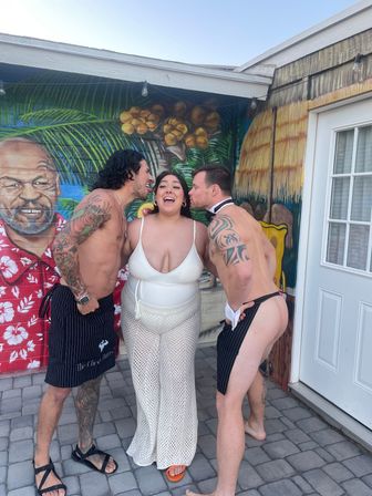 Laughing woman in a white swimsuit and crochet pants stands between two shirtless men in cheeky pinstripe aprons as they lean in to kiss her cheeks in front of a colorful tiki-style mural on a sunny patio.