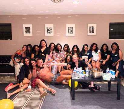 Fun indoor party group photo: about 15 women gathered in a living-room style space with a shirtless male entertainer reclining at the front, disco ball overhead, yellow balloons and a coffee table with bottled water and snacks.