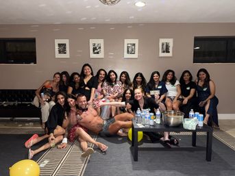 Fun indoor party group photo of about a dozen women on a couch and floor with a shirtless male entertainer posed across their laps, disco ball reflections, yellow balloons, scattered dollar bills, and a low table with bottled water and snacks.