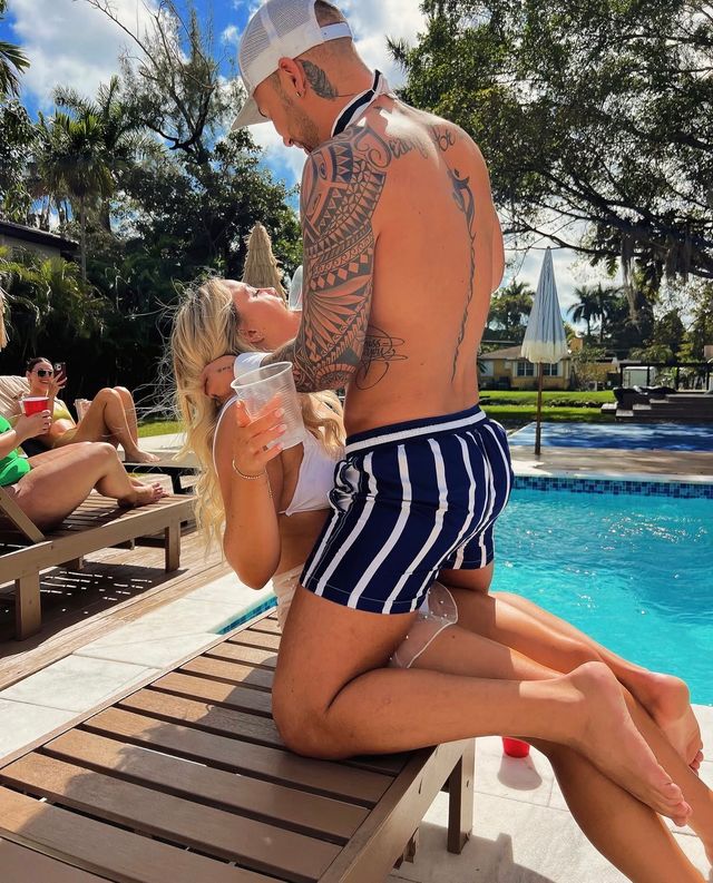 Poolside scene: tattooed man in striped swim trunks leans over a woman in a white bikini on a lounge chair, friends with drinks nearby at a sunny tropical outdoor pool.