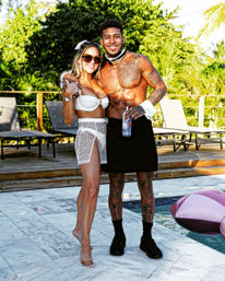 Two people in swimwear on a sunny tropical pool deck — woman in white bikini with sheer wrap and sunglasses, shirtless tattooed man in black skirt and cuffs holding a drink, lounge chairs and pink inflatable float nearby.