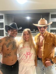 Three smiling people in a home kitchen at a bachelorette party in Palm Springs: woman in a white bachelorette tee between two men in cowboy hats—one shirtless with tattoos, one in a brown jacket and sunglasses.
