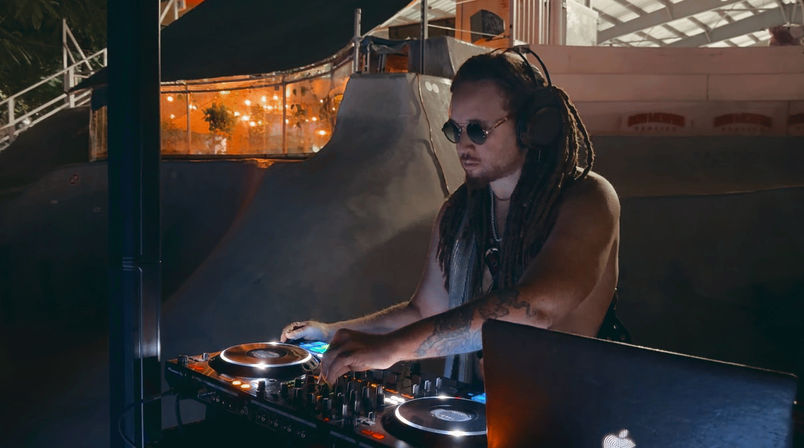 Nighttime outdoor DJ set at an urban skate bowl — a shirtless DJ with dreadlocks, sunglasses and headphones mixes on a glowing controller and laptop beneath warm string lights.