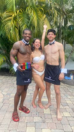 Three people smiling on a tropical poolside patio with palm trees: a woman in a white bikini raising her arm between two shirtless men in bow ties and striped aprons, one holding a rainbow set of shot glasses.