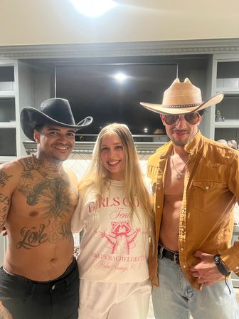 Smiling trio in a kitchen at a cowboy-themed bachelorette party: woman in a white bachelorette tee stands between two men in cowboy hats — one shirtless with chest tattoos, the other in an open tan shirt and sunglasses.