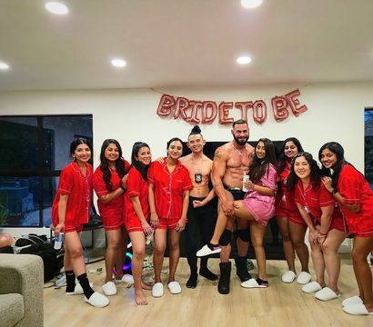Bachelorette party in a living room: bride-to-be in a pink robe and friends in matching red pajamas posing with two shirtless male performers under a 'BRIDE TO BE' balloon banner.