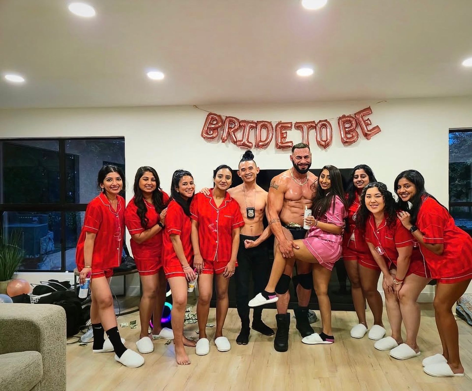 Bachelorette party in a living room: bride-to-be in a pink robe and friends in matching red pajamas posing with two shirtless male performers under a 'BRIDE TO BE' balloon banner.