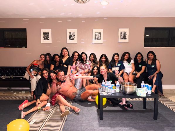 Lively indoor bachelorette party group photo: a dozen women on sofas and the floor posing with a shirtless male entertainer sprawled across laps, balloons and a coffee table with water bottles and snacks.