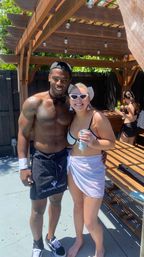 Two smiling adults at a sunny backyard pool party under a wooden pergola with string lights — shirtless server in a bow tie and apron beside a woman in white bikini, sunglasses and sarong holding a canned drink.