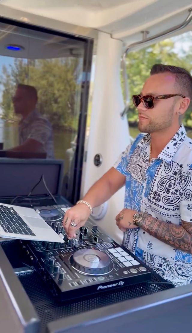 Tattooed DJ in sunglasses mixing on a laptop and DJ controller aboard a sunlit boat, with water and shoreline trees visible through a window.