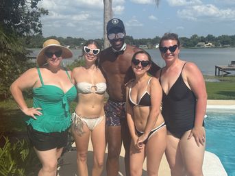 Five adults in swimsuits and sunglasses posing at a lakeside backyard pool with a palm tree and dock under a sunny summer sky.