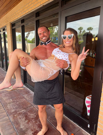 Two people at a sunny palm-lined patio: a muscular shirtless man in a bow tie and pinstripe apron lifts a smiling woman in heart-shaped sunglasses and a cherry-print bikini with a sheer skirt, both posing playfully by glass doors.
