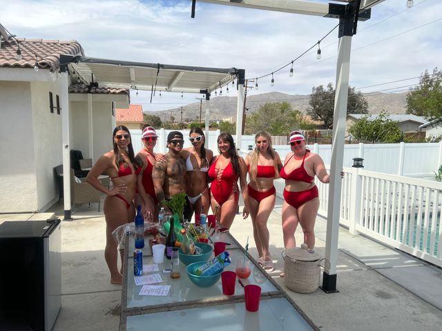 Group of friends in red swimsuits posing at a sunny backyard pool party on a patio with a pergola, drink-filled table, and desert mountains in the distance.