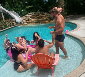 Adults enjoying a sunny backyard pool party: friends in swimwear lounging on pink inflatables and a shallow ledge, sipping drinks near a waterslide and rock waterfall surrounded by tropical plants.