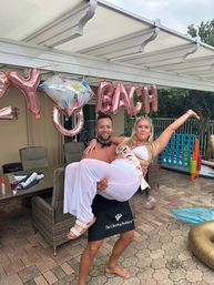 Poolside patio bachelorette party — shirtless man in a black apron lifts a woman in white with a bride-to-be sash under rose-gold balloons, wicker furniture and a giant outdoor game nearby.