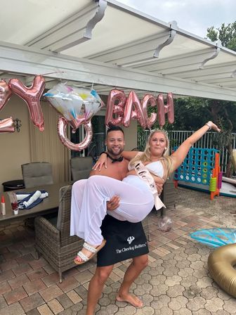 Poolside patio bachelorette party — shirtless man in a black apron lifts a woman in white with a bride-to-be sash under rose-gold balloons, wicker furniture and a giant outdoor game nearby.